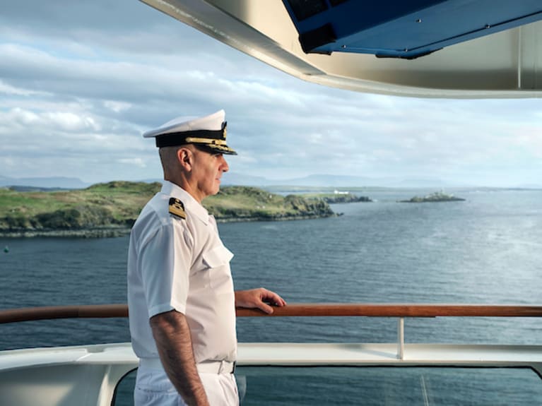 Misty sky hints at rain as captain stands confidently by the ship's balustrade. The side angle shot captures the allure of seafaring adventure.
