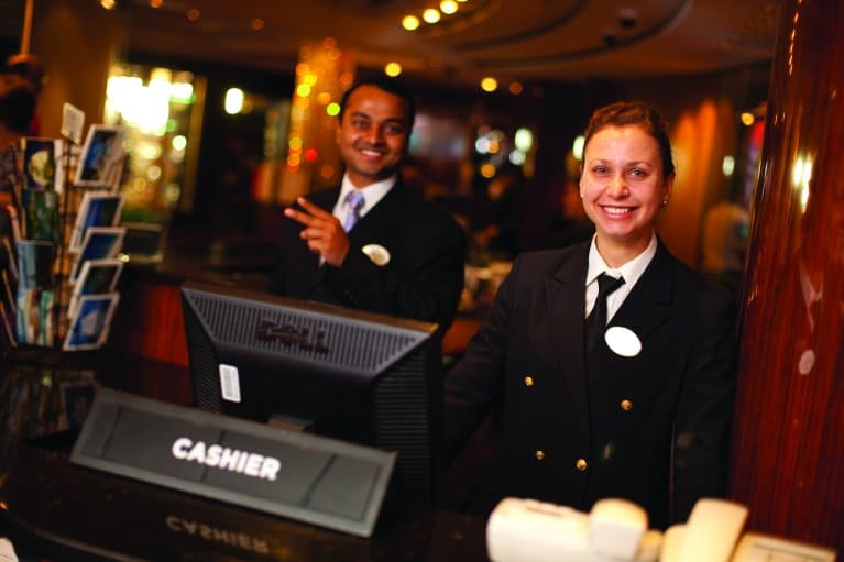 Two crew members in clean black uniforms stand at a polished guest services counter on a Norwegian Cruise ship, smiling and ready to assist guests.