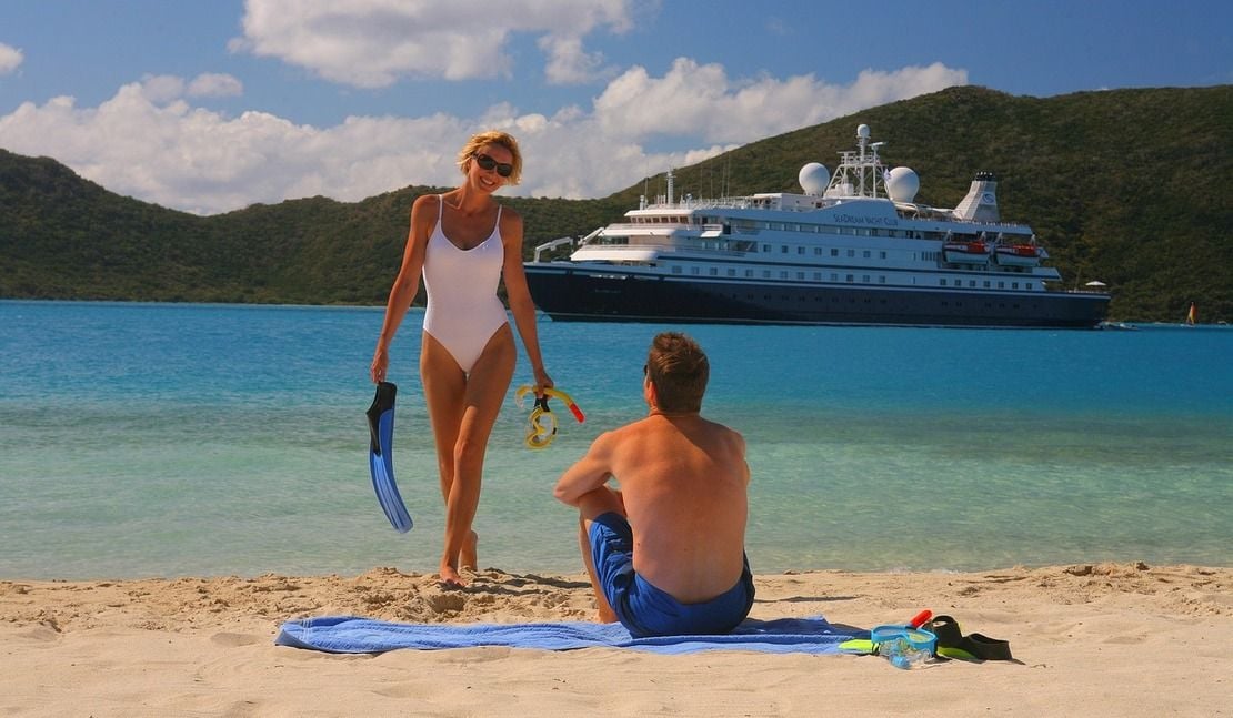 Lady coming out of the water to a man sitting on a beach in the Caribbean with a Seadream Yacht club ship in the background - supporting the article on The Ultimate Guide to Choosing your Perfect Cruise Holiday